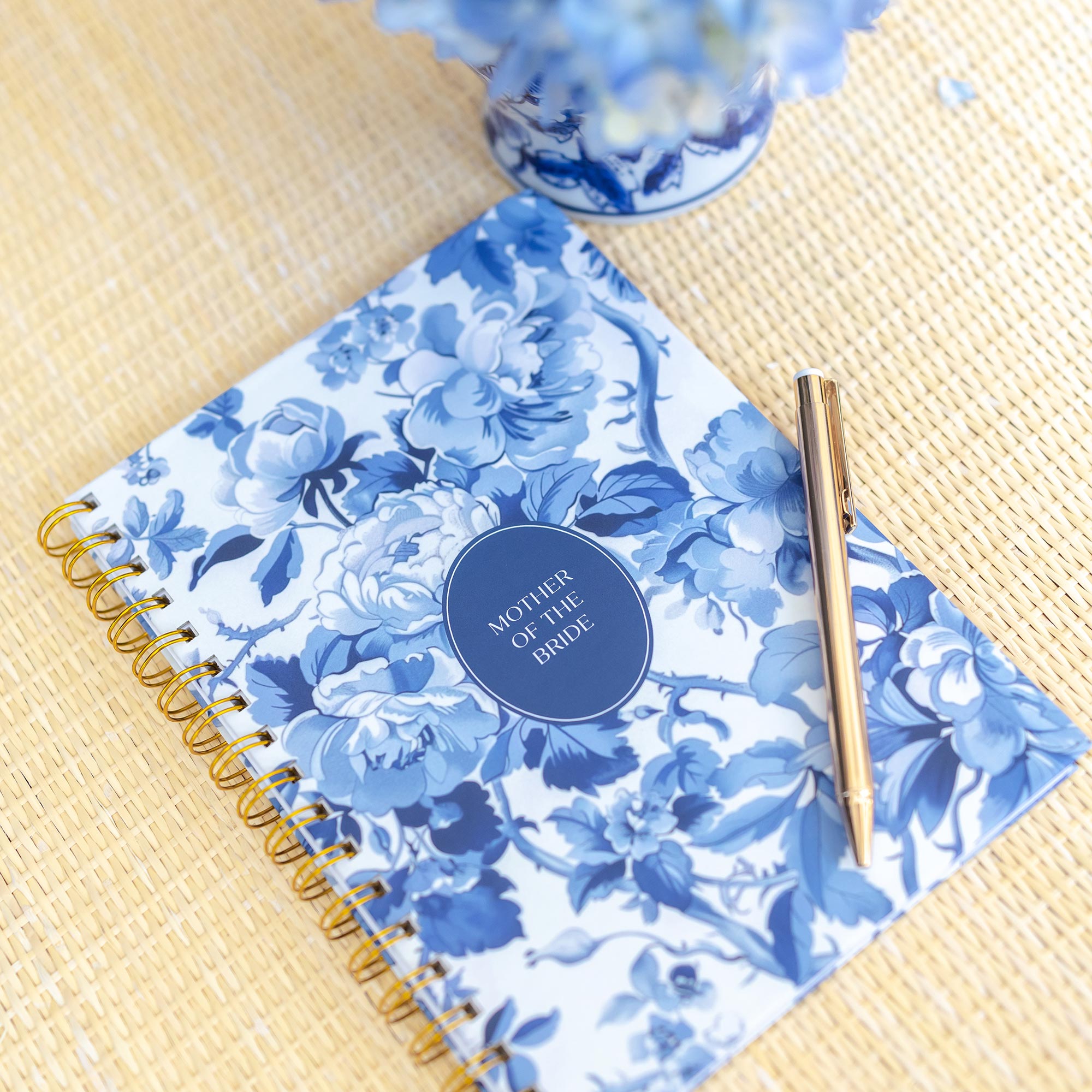 Styled flatlay featuring a Keepsake Paper Co. blue toile mother of the bride planner on a rattan surface