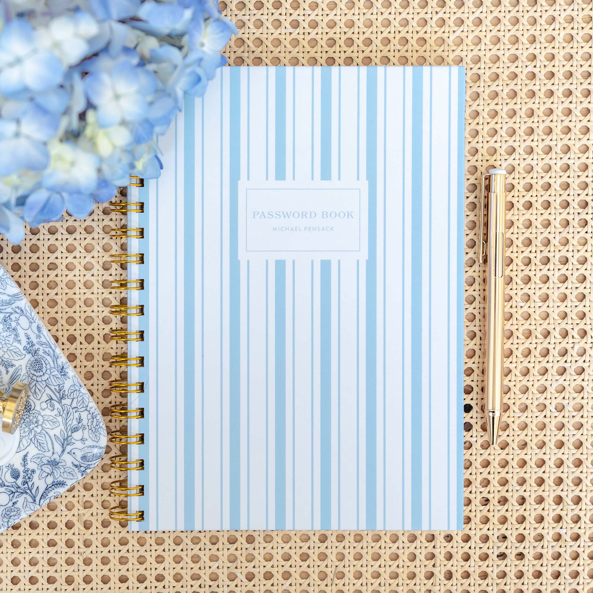 Styled flatlay featuring a Keepsake Paper Co. blue and white stripe password book on a rattan background with hydrangeas