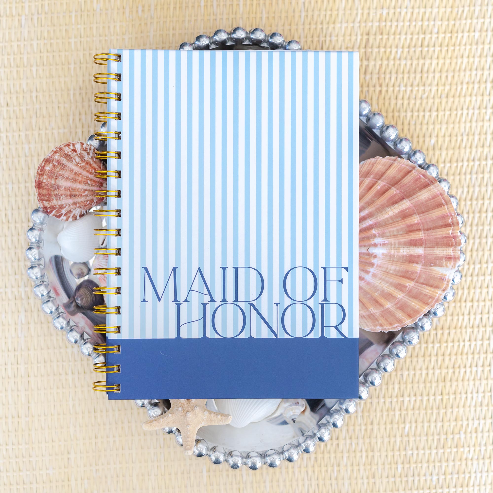 Styled flatlay featuring a Keepsake Paper Co. classic stripe maid of honor planner on top of seashells in a silver tray on a rattan background