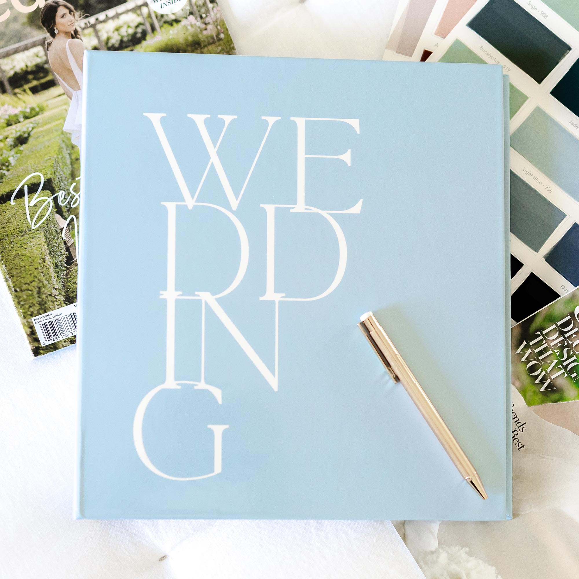 Styled flatlay featuring a Keepsake Paper Co. minimalist wedding planning binder on top of fabric swatches and wedding magazines with a gold pen