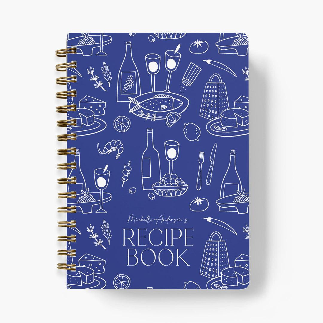 Recipe book with blue cover featuring white Mediterranean food illustrations and gold spiral binding