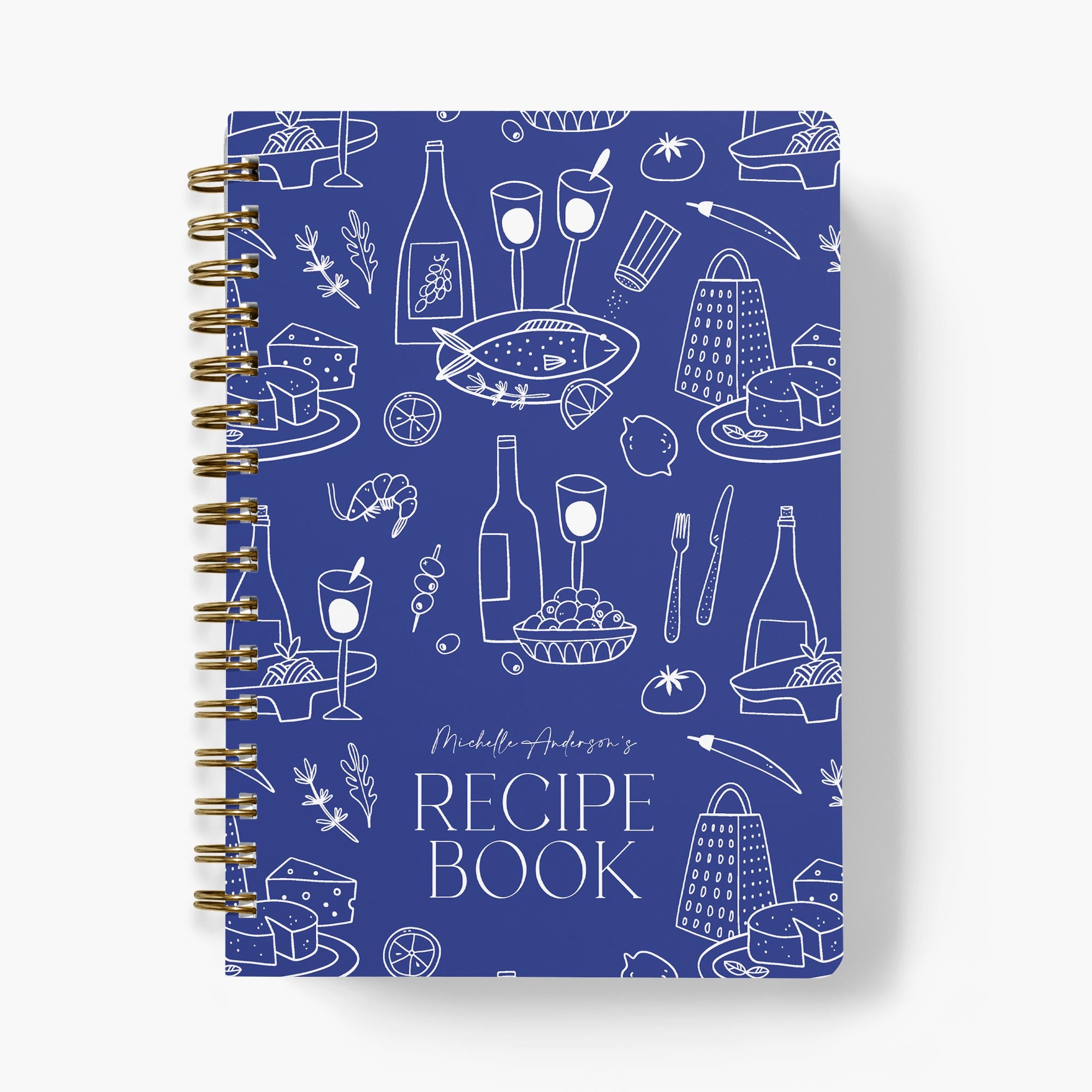 Recipe book with blue cover featuring white Mediterranean food illustrations and gold spiral binding