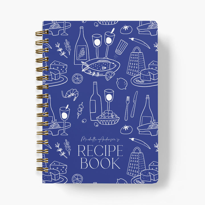Recipe book with blue cover featuring white Mediterranean food illustrations and gold spiral binding