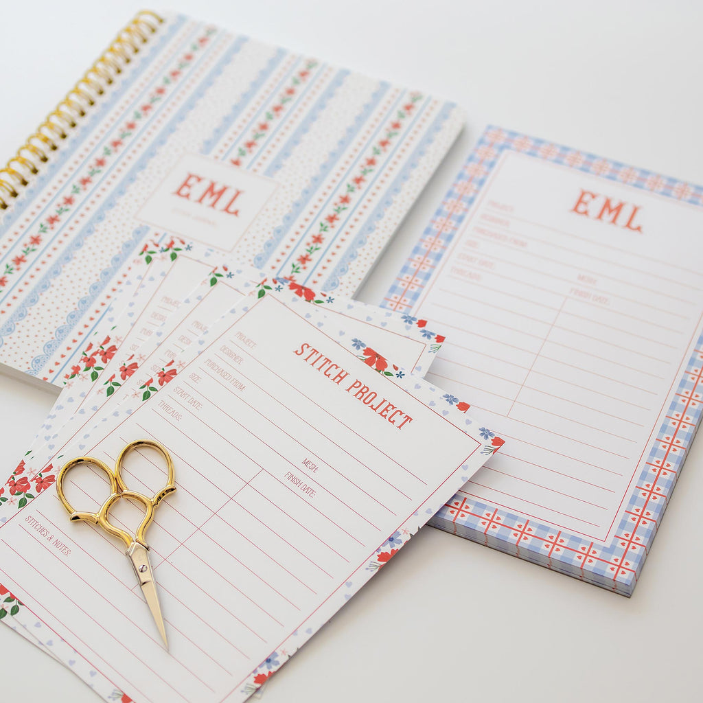 Needlepoint project products such as a journal, notepad and stitch project cards styled with gold scissors on a white background