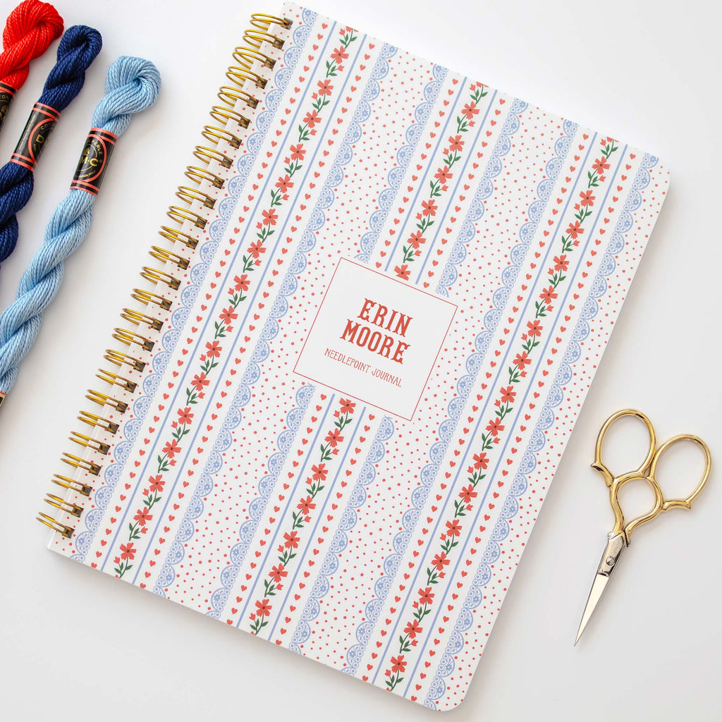 Needlepoint notebook with heart and floral pattern surrounded by thread and scissors on a white background