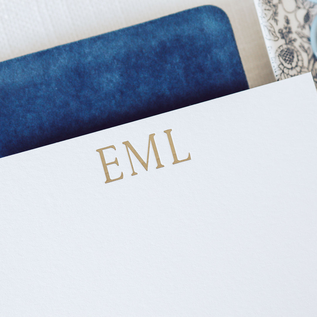 A personalized monogram notecard printed in gold foil on white cotton paper sits on top of a blue notebook