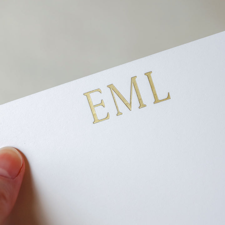 A hand holds a custom gold foil notecard featuring a 3 letter monogram over a neutral background