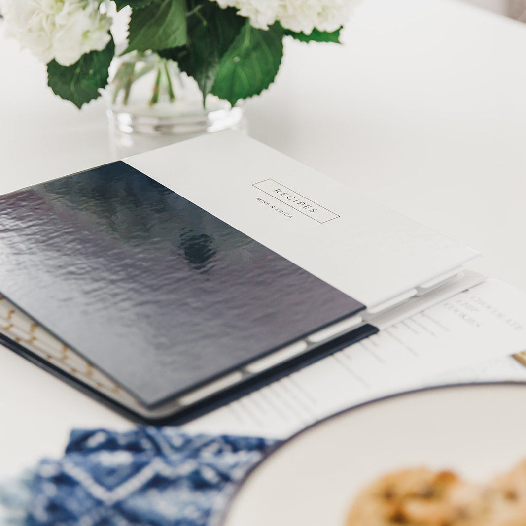 Personalized navy blue recipe binder with gold foil lettering reading 'RECIPES, MIKE & ERICA' resting on a wooden table beside a green potted plant