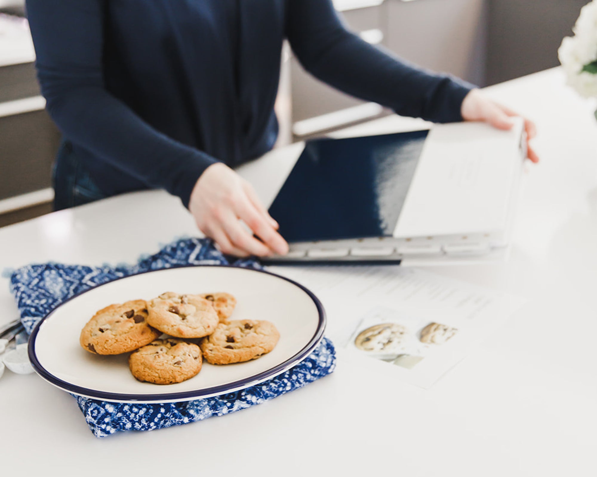 Person reaching for a Keepsake Paper Co. recipe binder on a white shelf, with homemade cookies and folded blue linen in the foreground