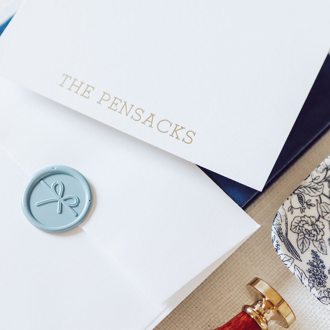 A personalized notecard and envelope printed in gold foil on white cotton paper styled with blue accessories